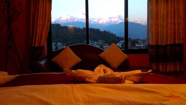 A hotel room with a bed, two pillows, a window with a mountain view, and curtains. - Hotel Orchid (Pokhara)