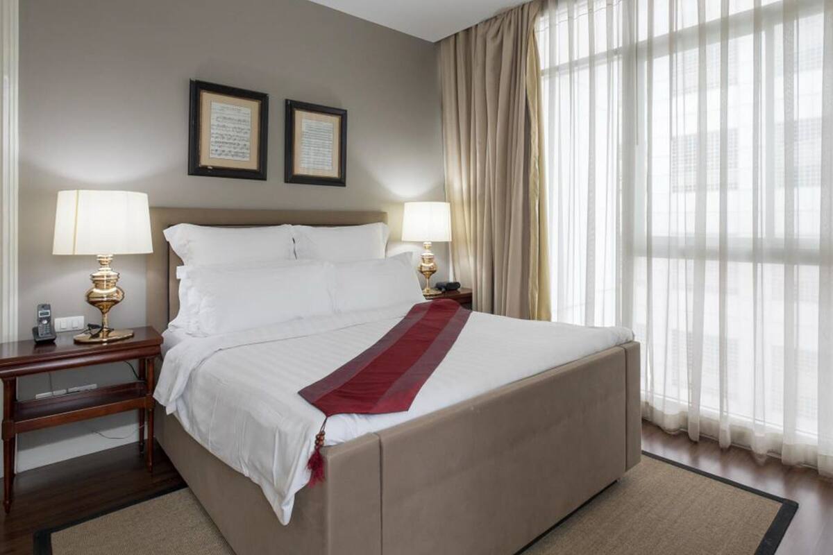 Ein Hotelzimmer mit einem großen Bett, zwei Nachttischlampen, einem Nachttisch und einem Fenster mit durchsichtigen Vorhängen.