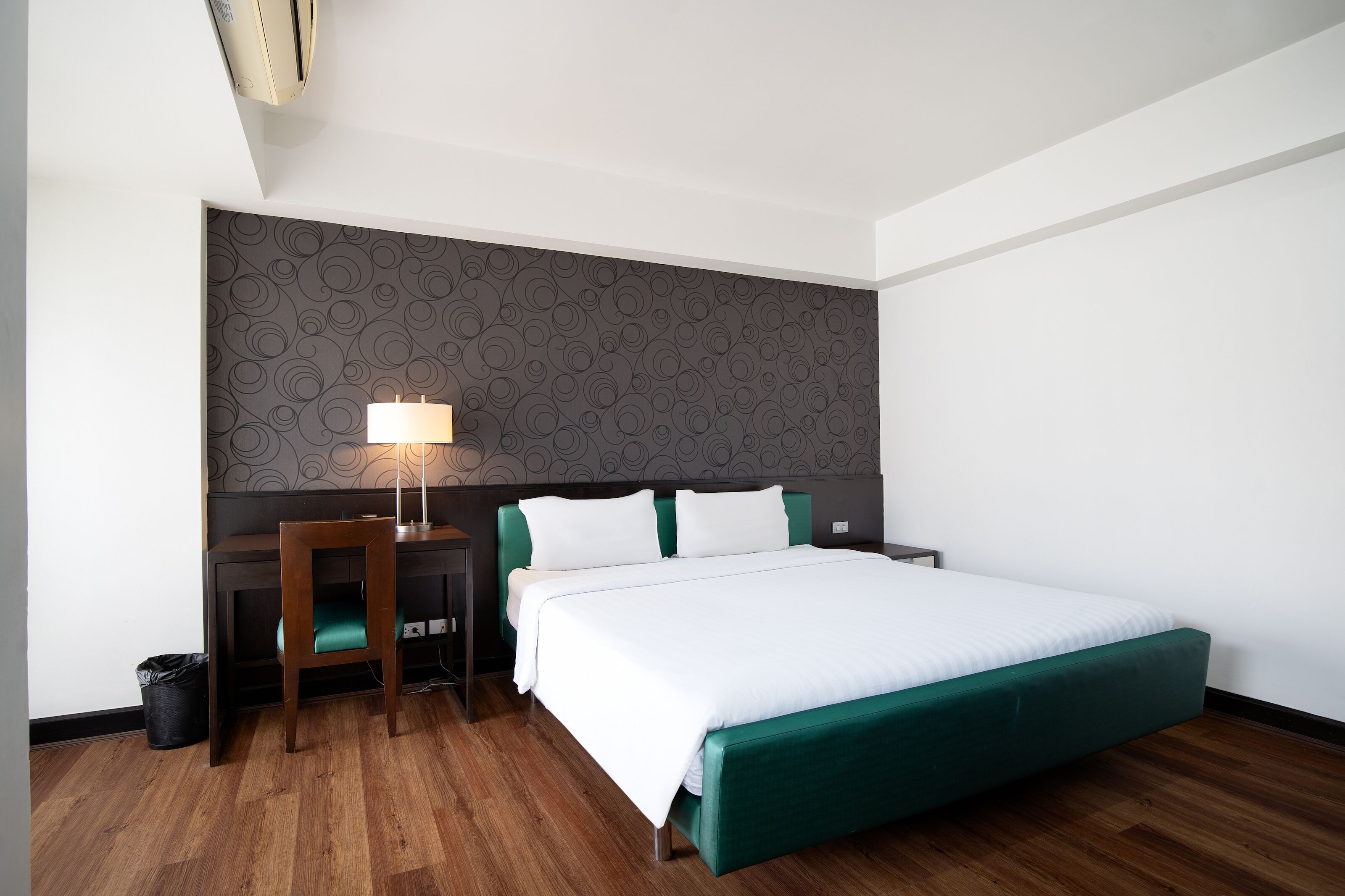 A hotel room with a bed, a desk, a chair, a lamp, and a patterned wall.