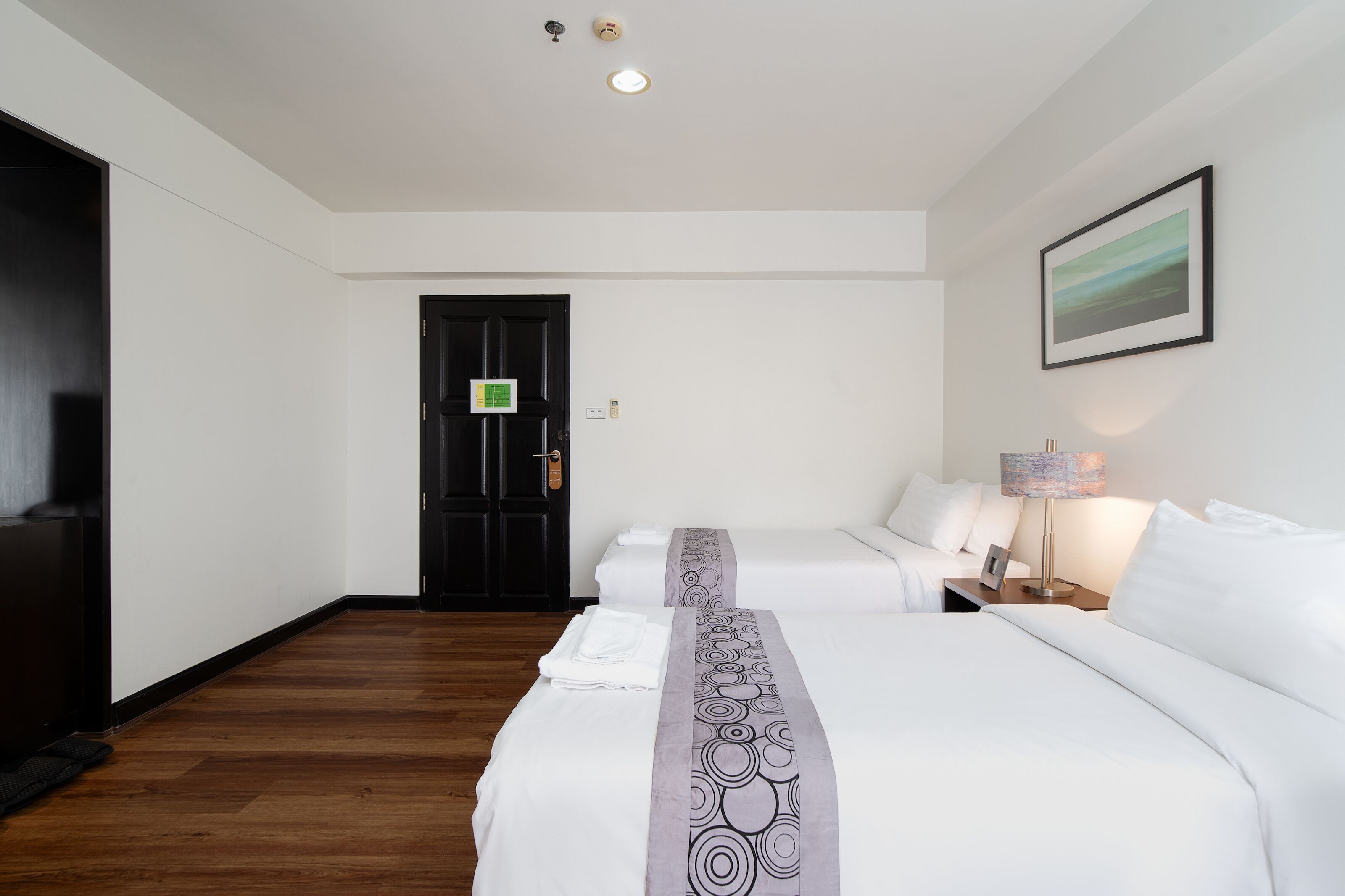 Ein Hotelzimmer mit zwei Betten, Holzboden, einer schwarzen Tür, einem gerahmten Bild an der Wand und einer Nachttischlampe.