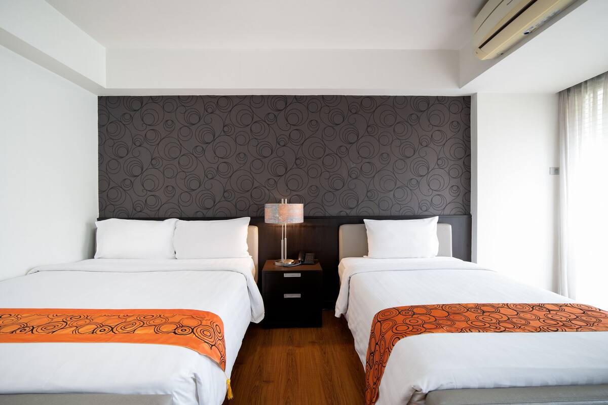 Hotellihuone, jossa on kaksi sänkyä, yöpöytä lampun kanssa, kuvioitu pääty ja ikkuna, jossa on verhot.