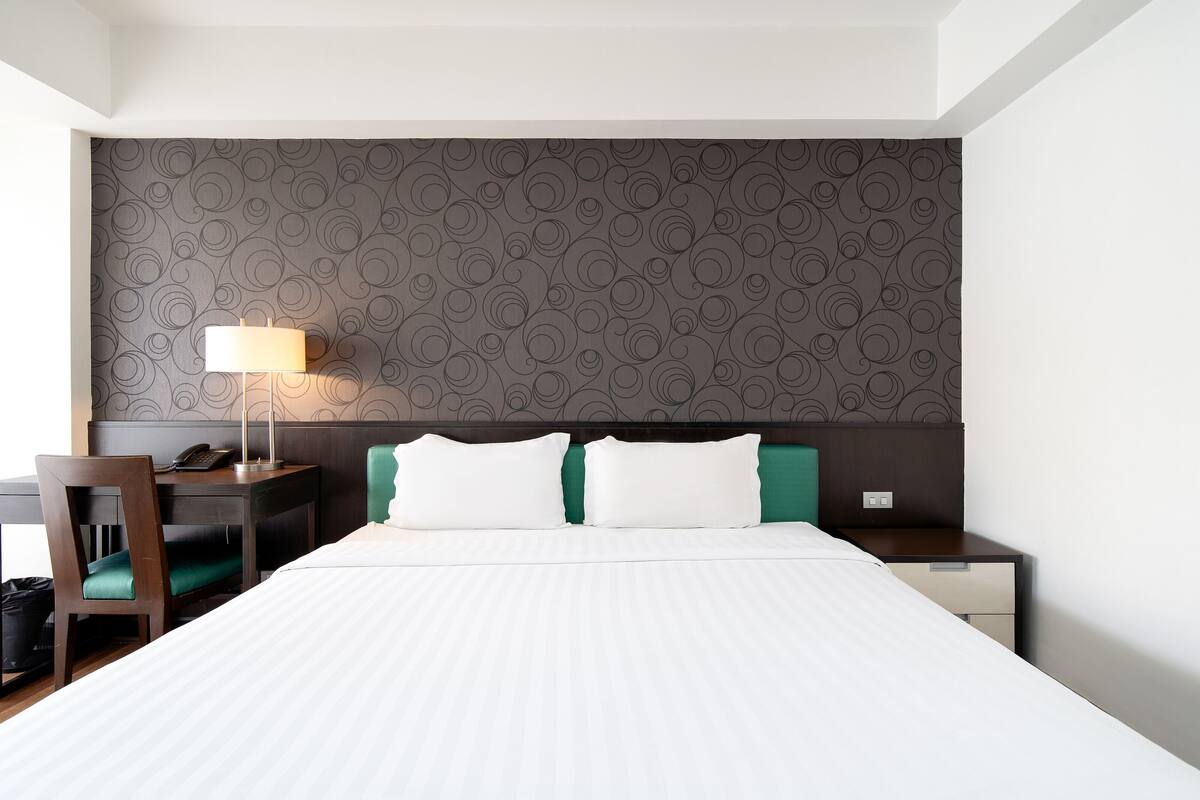 Hotellihuone, jossa on suuri sänky, työpöytä lampun kanssa, tuoli ja yöpöytä.