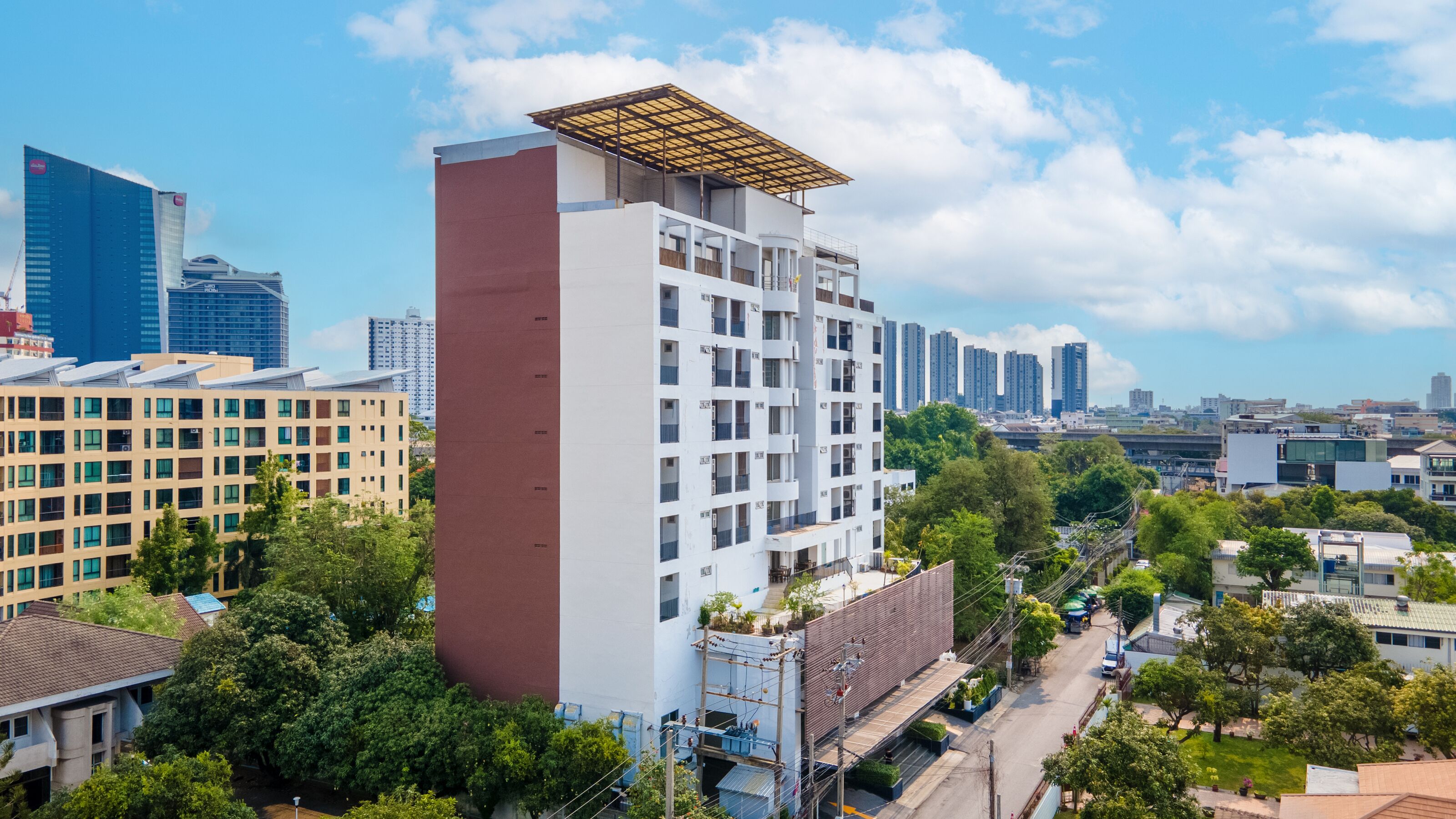 A modern high-rise building with a flat roof and multiple balconies, surrounded by greenery and other buildings.