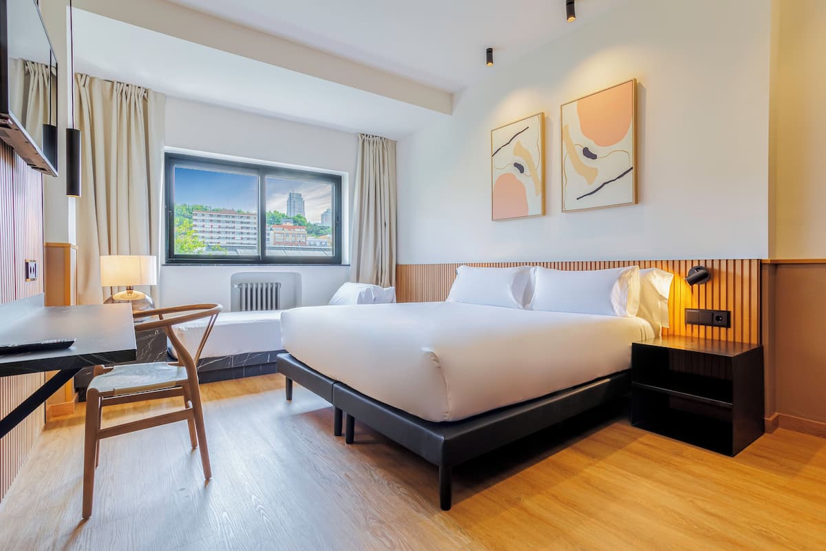 A modern hotel room with a bed, desk, chair, and a view of the city through a large window.