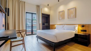 A hotel room with a bed, a desk with a laptop, a chair, a nightstand, and a view of the city through a large window. - Hotel Acta Madfor (Madrid)