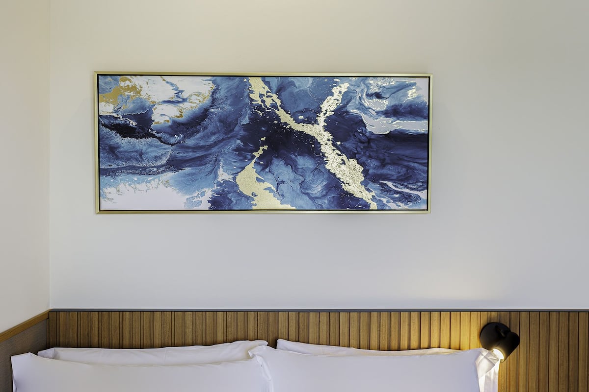 A room with a bed featuring a wooden headboard, a large abstract painting on the wall, and a wall-mounted light fixture.