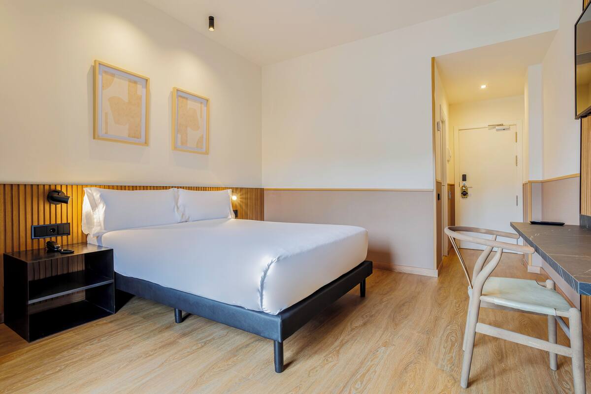 A modern hotel room with a large bed, bedside table, and a desk area.
