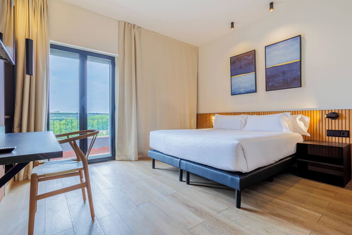 A hotel room with a bed, a desk, a chair, a TV, and a balcony with curtains.