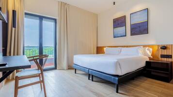 Habitación de hotel con cama, escritorio, silla, televisor y balcón con cortinas.