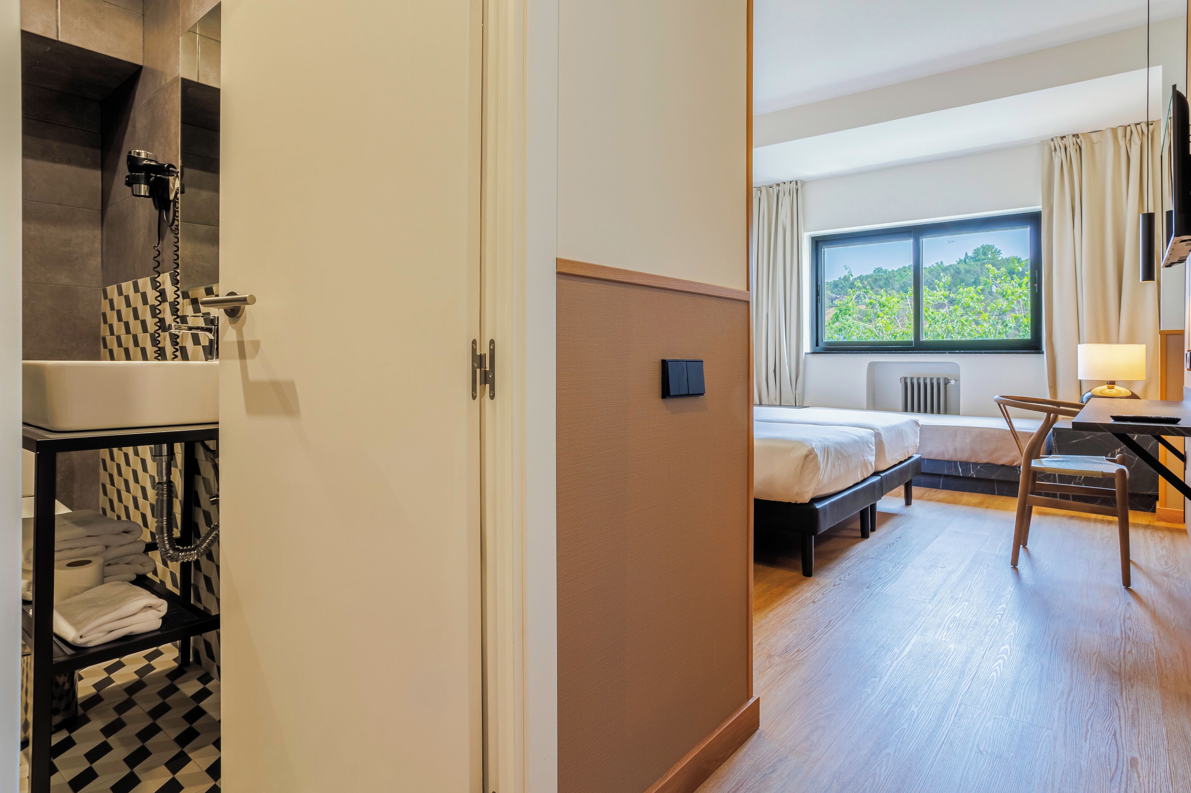 Una habitación de hotel con piso a cuadros, una cama, un escritorio con silla, un ventanal con vista a follaje y un perchero con toallas.