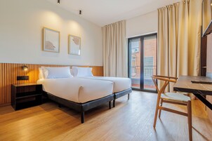 Una habitación de hotel moderna con una cama doble, una mesita de noche de madera, un escritorio con silla y ventanas grandes con cortinas.