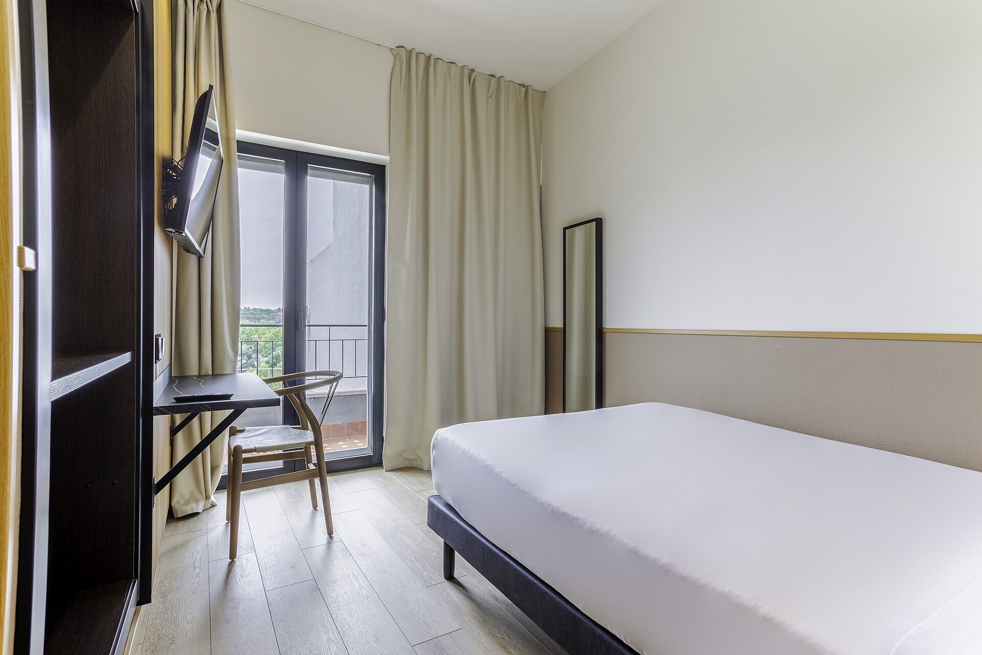Habitación de hotel con cama, escritorio, silla, televisor y balcón con cortinas.