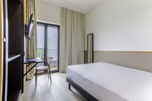 Habitación de hotel con cama, escritorio, silla, televisor y balcón con cortinas.