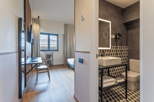 Una habitación de hotel moderna con un escritorio de madera, una cama, un baño con suelo a cuadros blanco y negro y un ventanal con cortinas.
