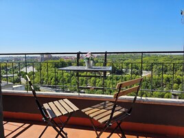 Un balcón con mesa y sillas, con vistas a un paisaje urbano con zonas verdes y edificios.