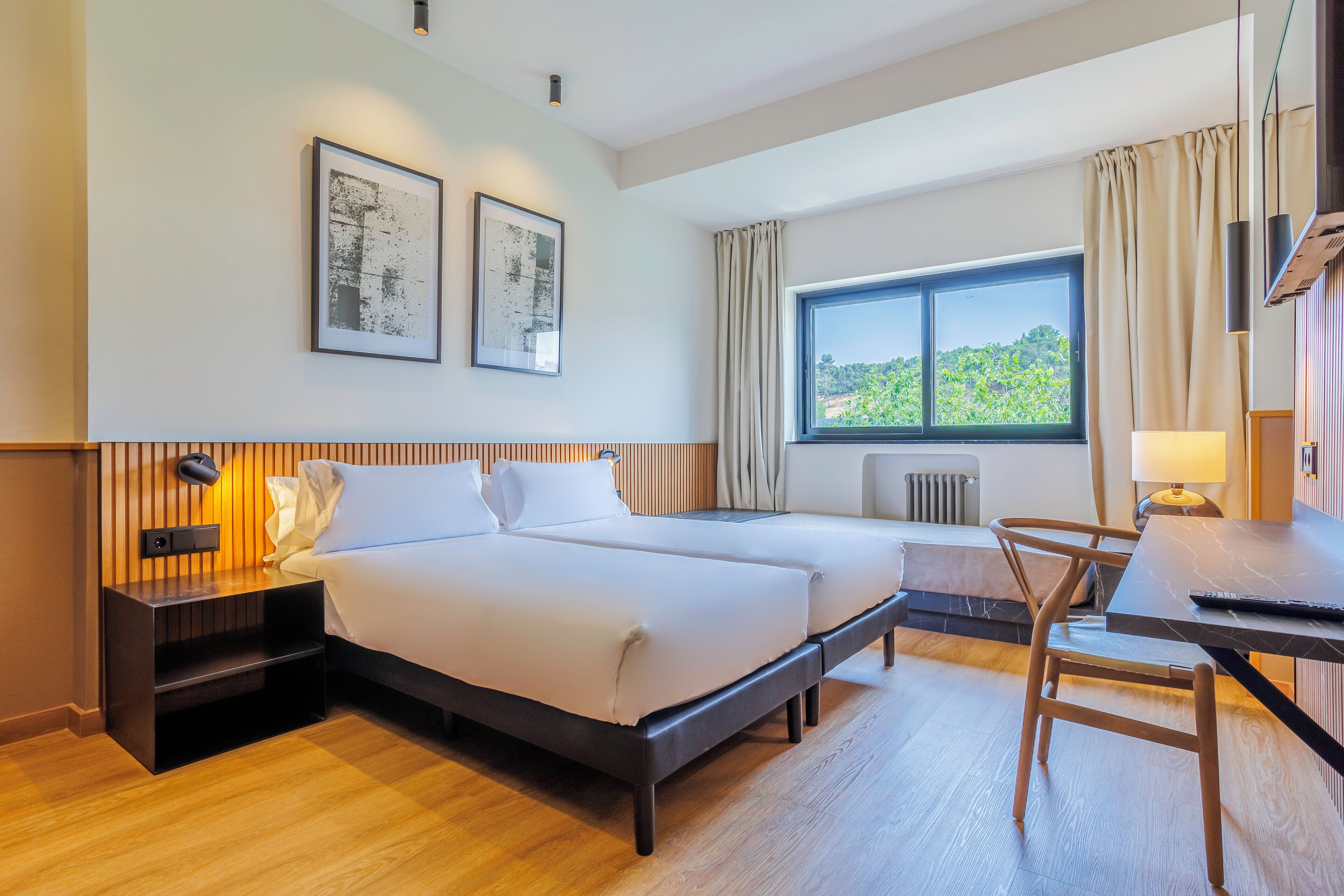 Habitación de hotel con cama, mesitas de noche, un escritorio y ventana con vista a zonas verdes.