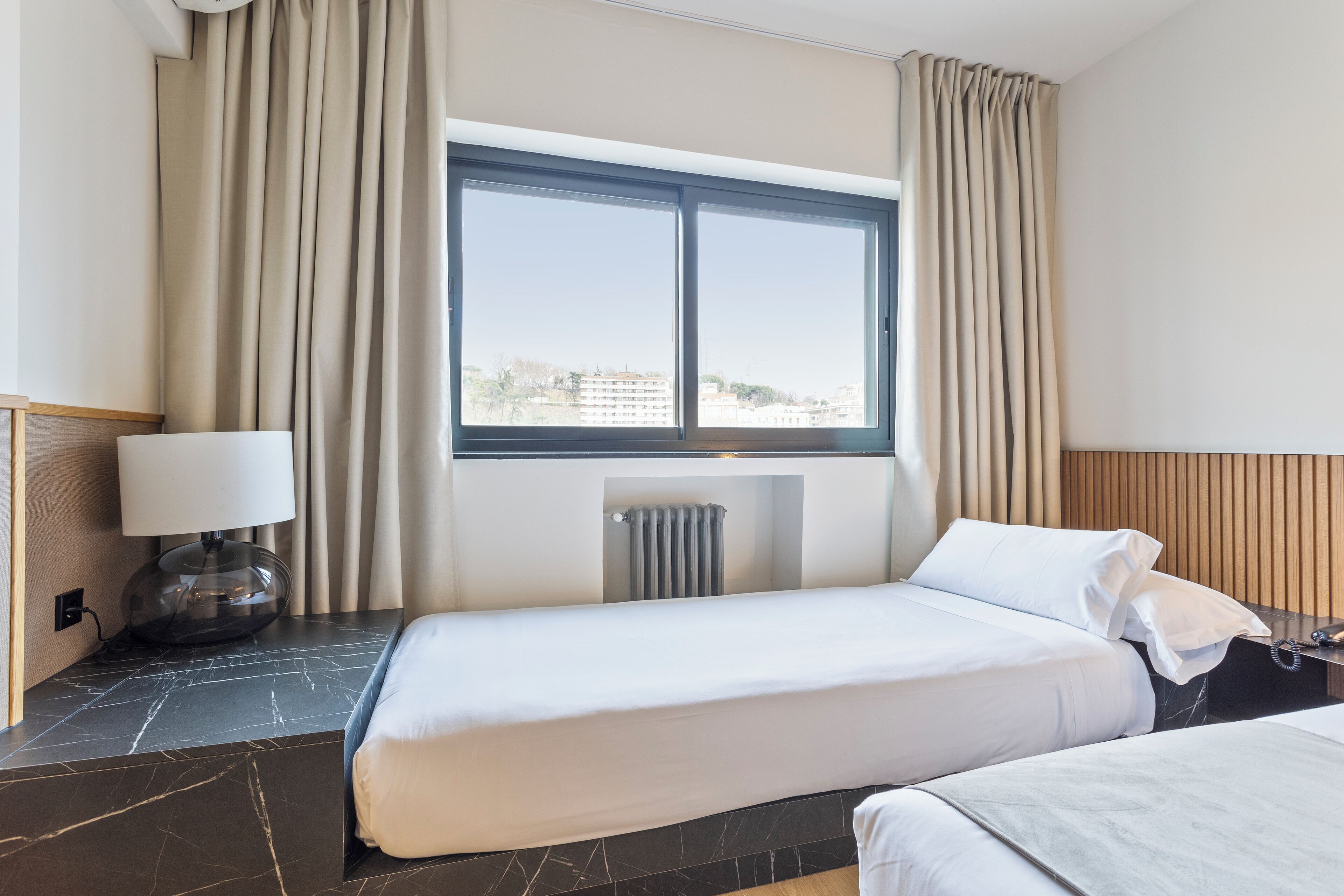 A hotel room with two beds, a window with curtains, a lamp, and a radiator.