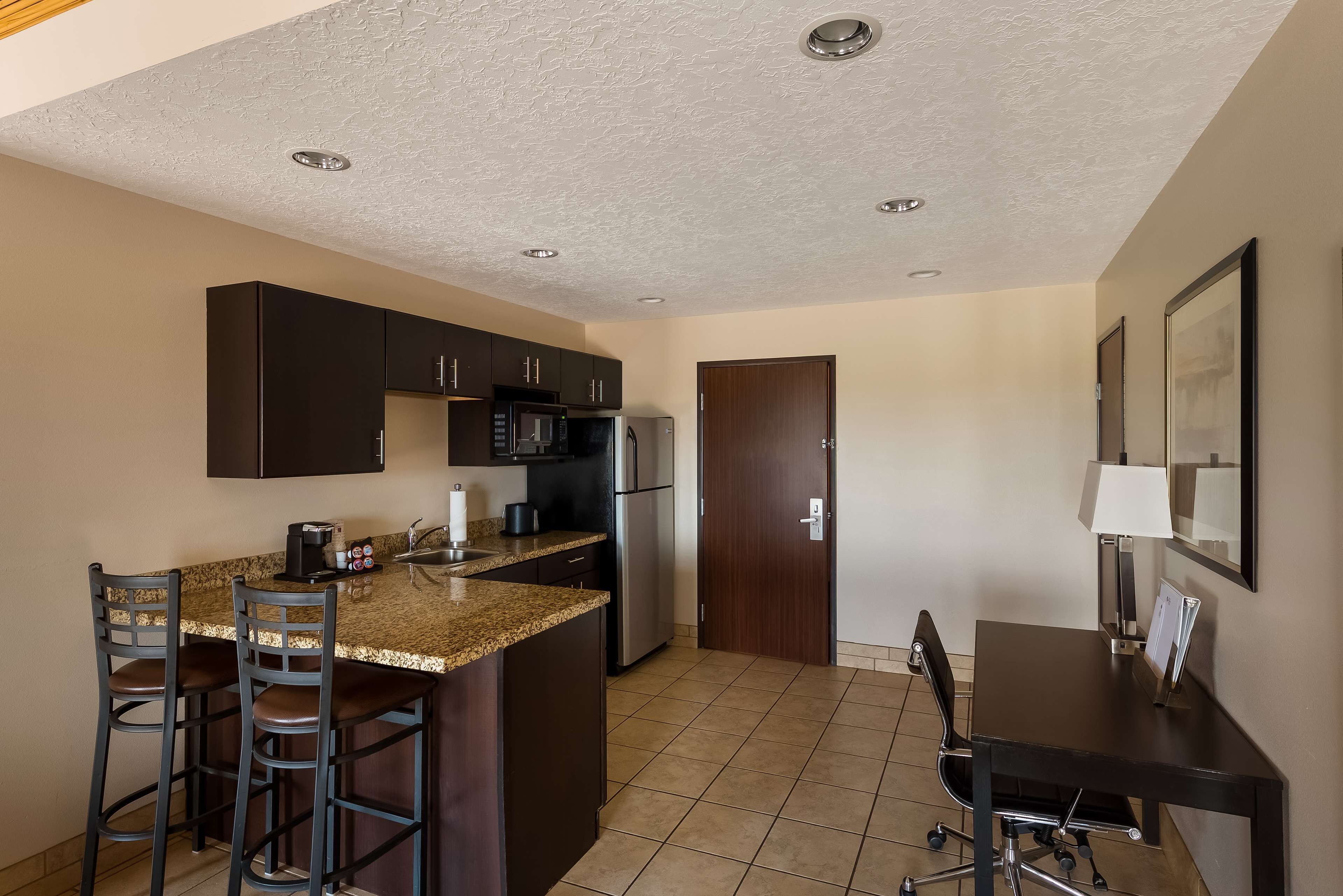 A compact kitchen with dark cabinets, a granite countertop, a microwave, a refrigerator, and a small island with bar stools.