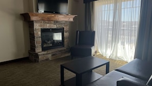 A living room with a stone fireplace, a flat-screen TV mounted above it, a leather armchair, and a large window with sheer curtains. - Best Western Plus Landmark Hotel (Roosevelt)