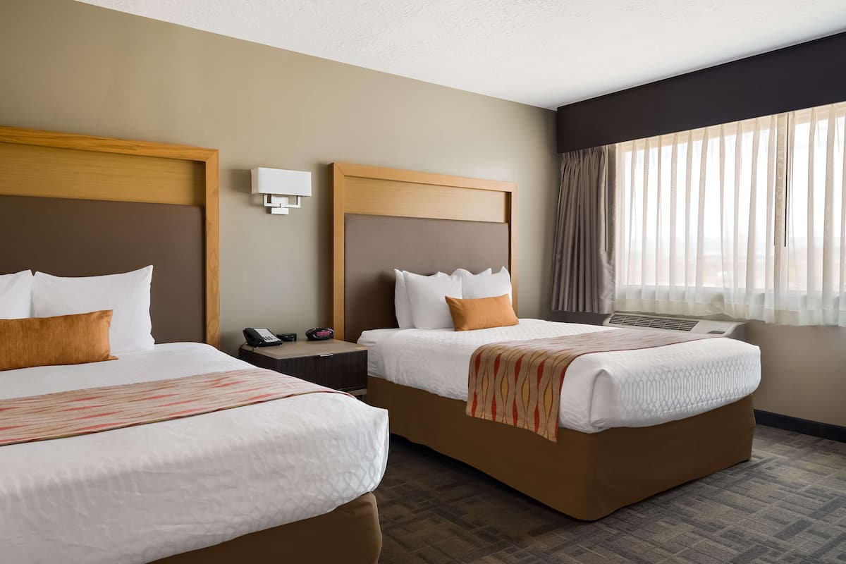 A hotel room with two beds, a bedside table with a phone, a wall-mounted lamp, and a window with sheer curtains.