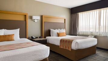 A hotel room with two beds, a bedside table with a phone, a wall-mounted lamp, and a window with sheer curtains.