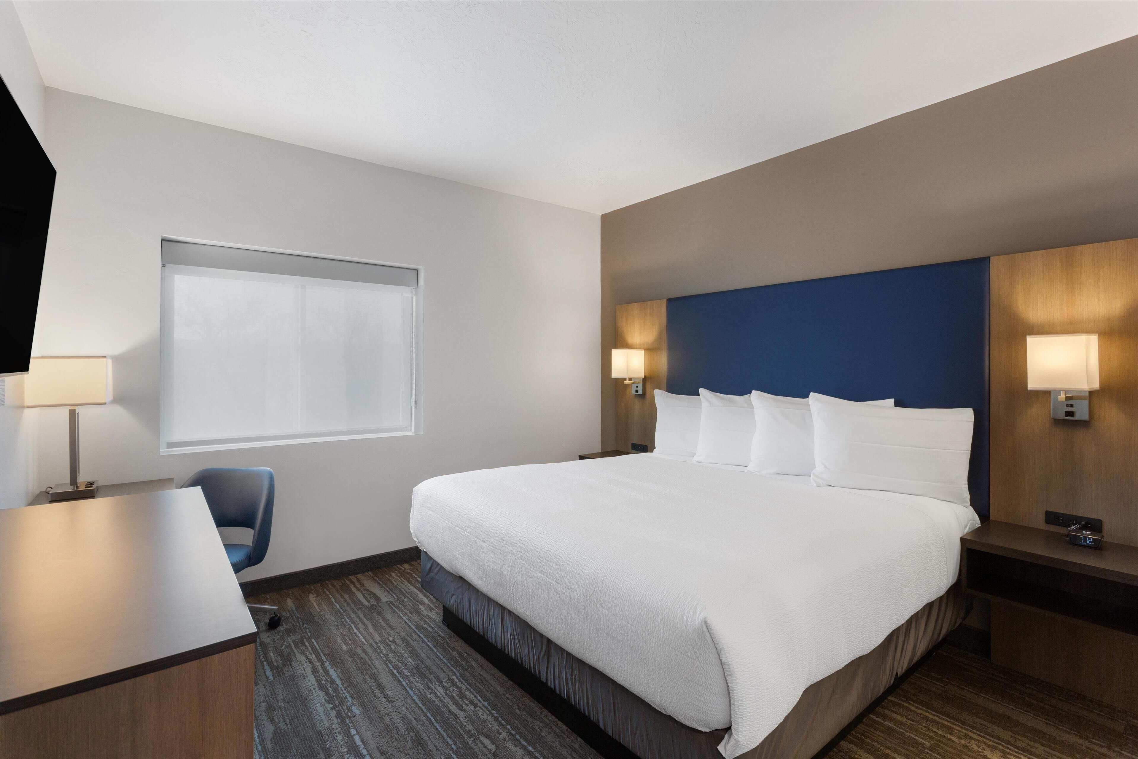A hotel room with a large bed, a desk, a chair, and a window with blinds.