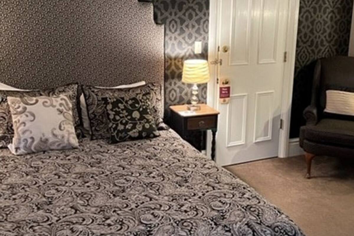 A bedroom with a patterned bedspread, a nightstand with a lamp, a chair, and a wardrobe.