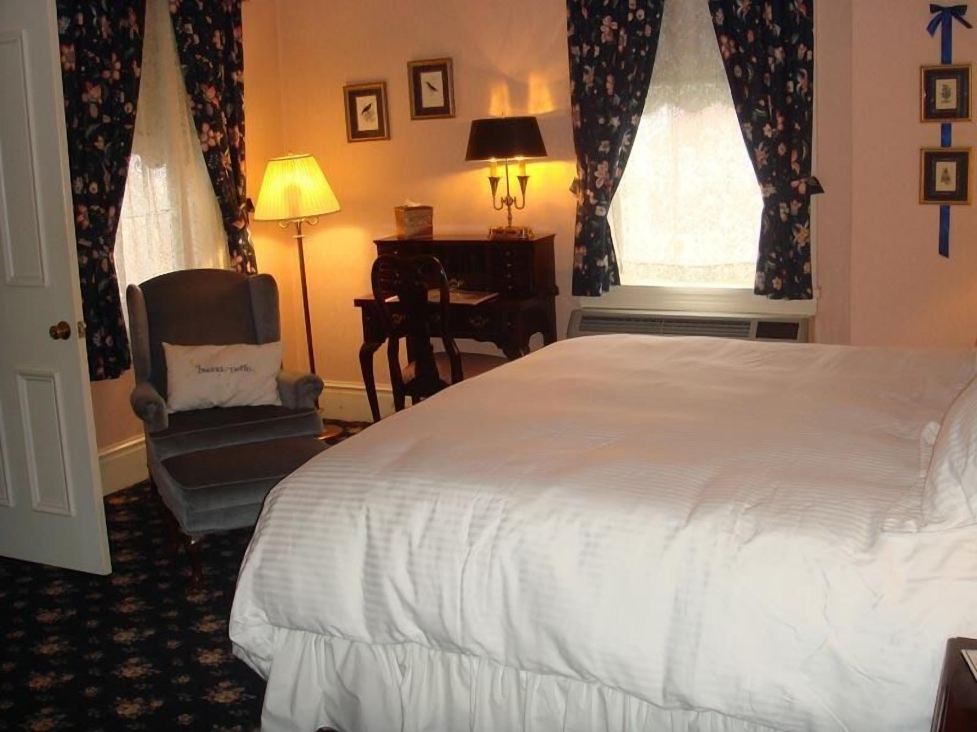 A hotel room with a large bed, a desk with a chair, two lamps, and a window with curtains.