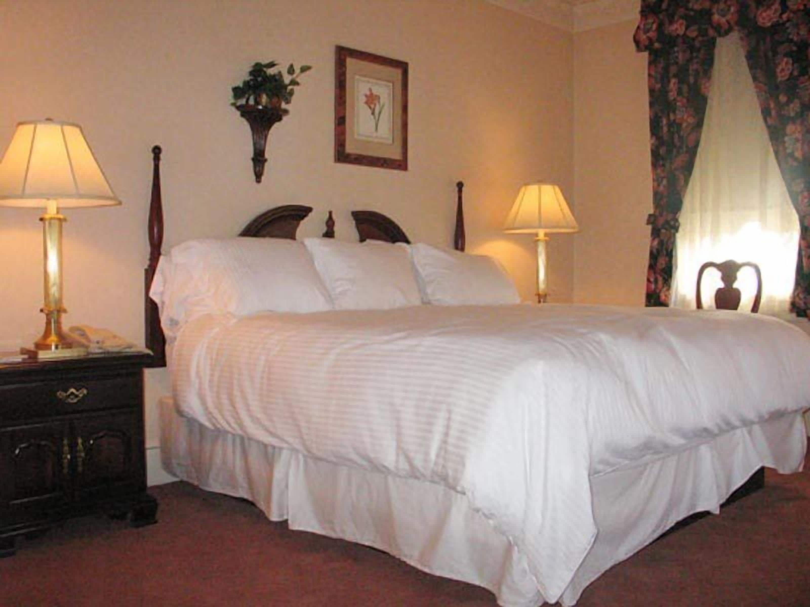 A neatly made bed with white linens, two bedside lamps, a dark wooden nightstand, a framed picture on the wall, and a floral curtain.