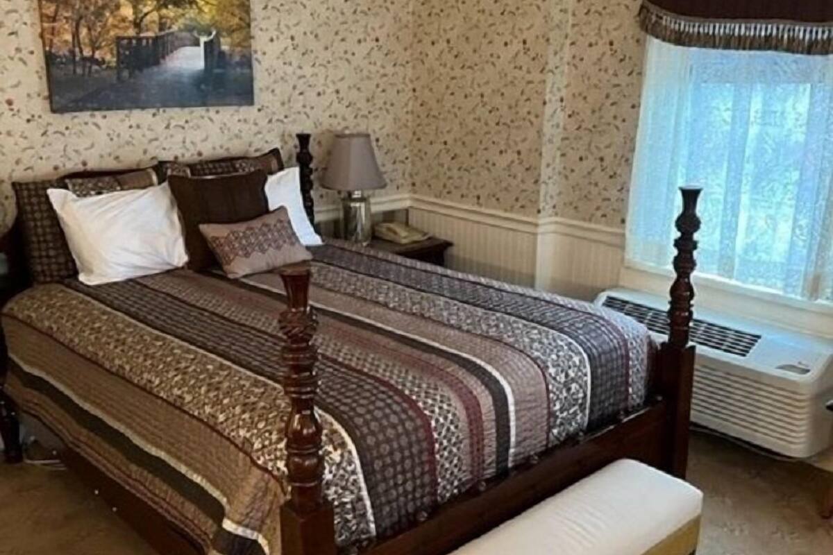 A bedroom with a wooden four-poster bed, a bench, a nightstand, a lamp, and a window with a curtain.