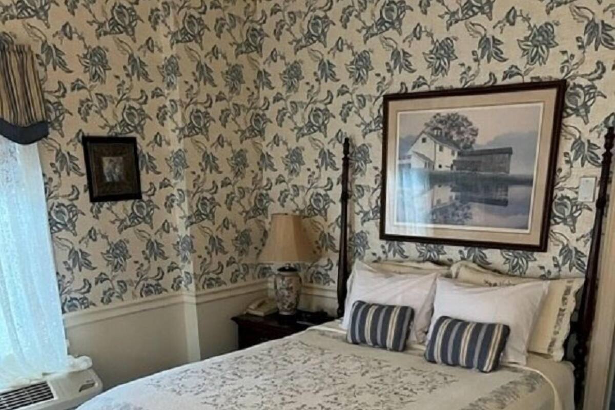 A bedroom with a large bed, floral wallpaper, a framed picture on the wall, and a bedside table with a lamp.