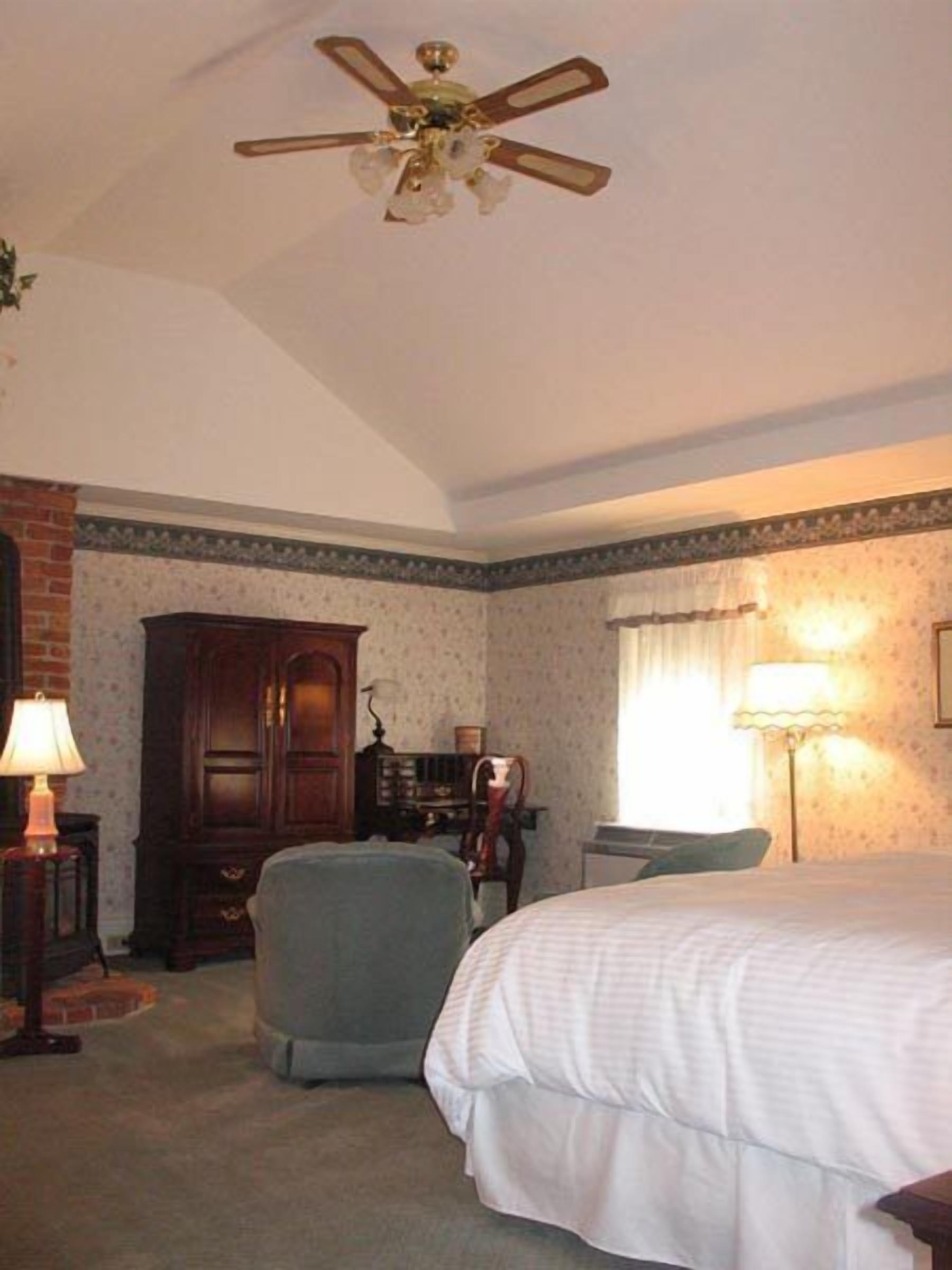 Habitación de hotel con cama, una silla, un escritorio, una lámpara, ventilador de techo y pared de ladrillo.