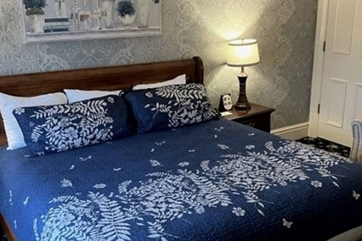 A bedroom with a wooden bed, floral-patterned bedding, a bedside lamp, and a chair.