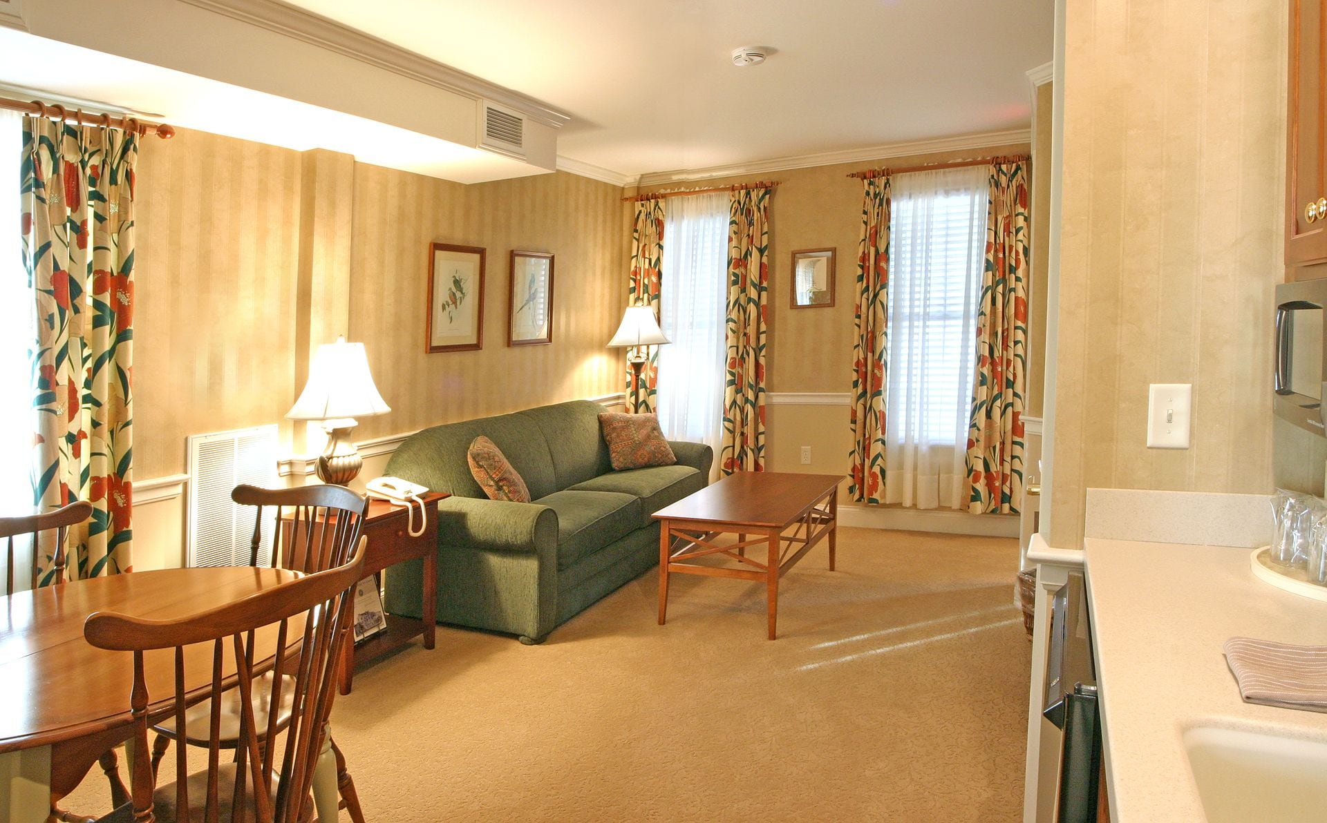 A living room with a green sofa, wooden coffee table, and a dining table with chairs. There are floral curtains on the windows, and a lamp on the coffee table.
