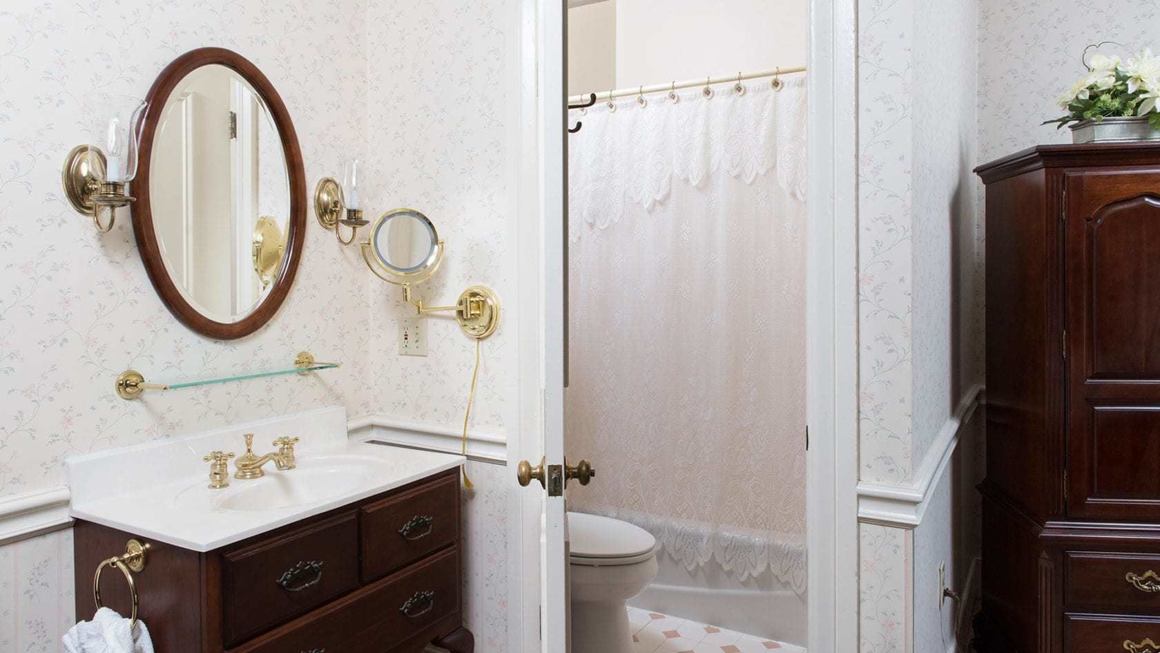Un baño con un lavabo blanco, un espejo, un espejo redondo pequeño, una cortina de ducha de vidrio y un armario de madera.