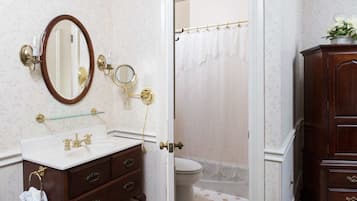 Un baño con un lavabo blanco, un espejo, un espejo redondo pequeño, una cortina de ducha de vidrio y un armario de madera.