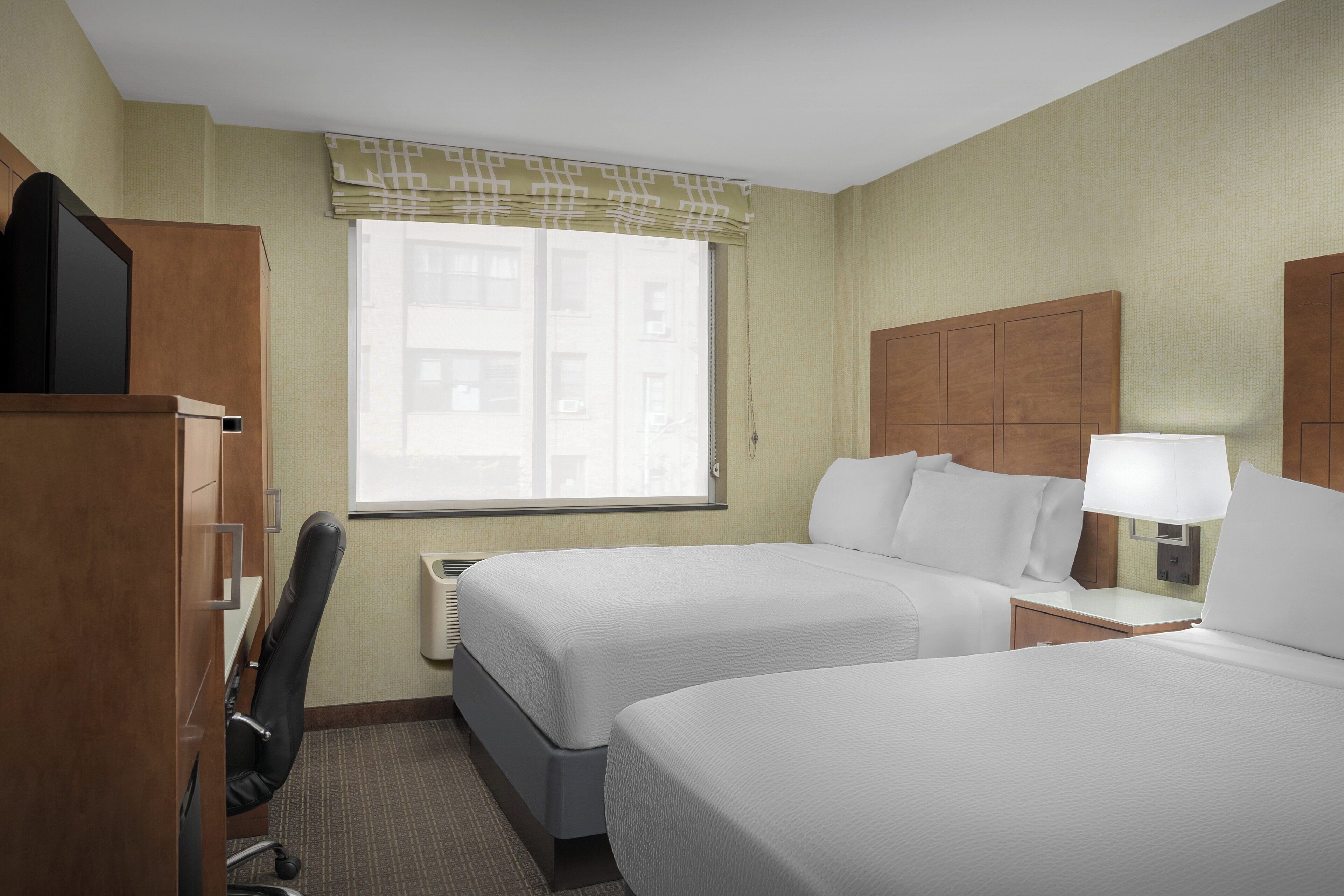 Quarto de hotel com duas camas, uma escrivaninha, uma cadeira, uma televisão e uma janela com vista para edifícios.