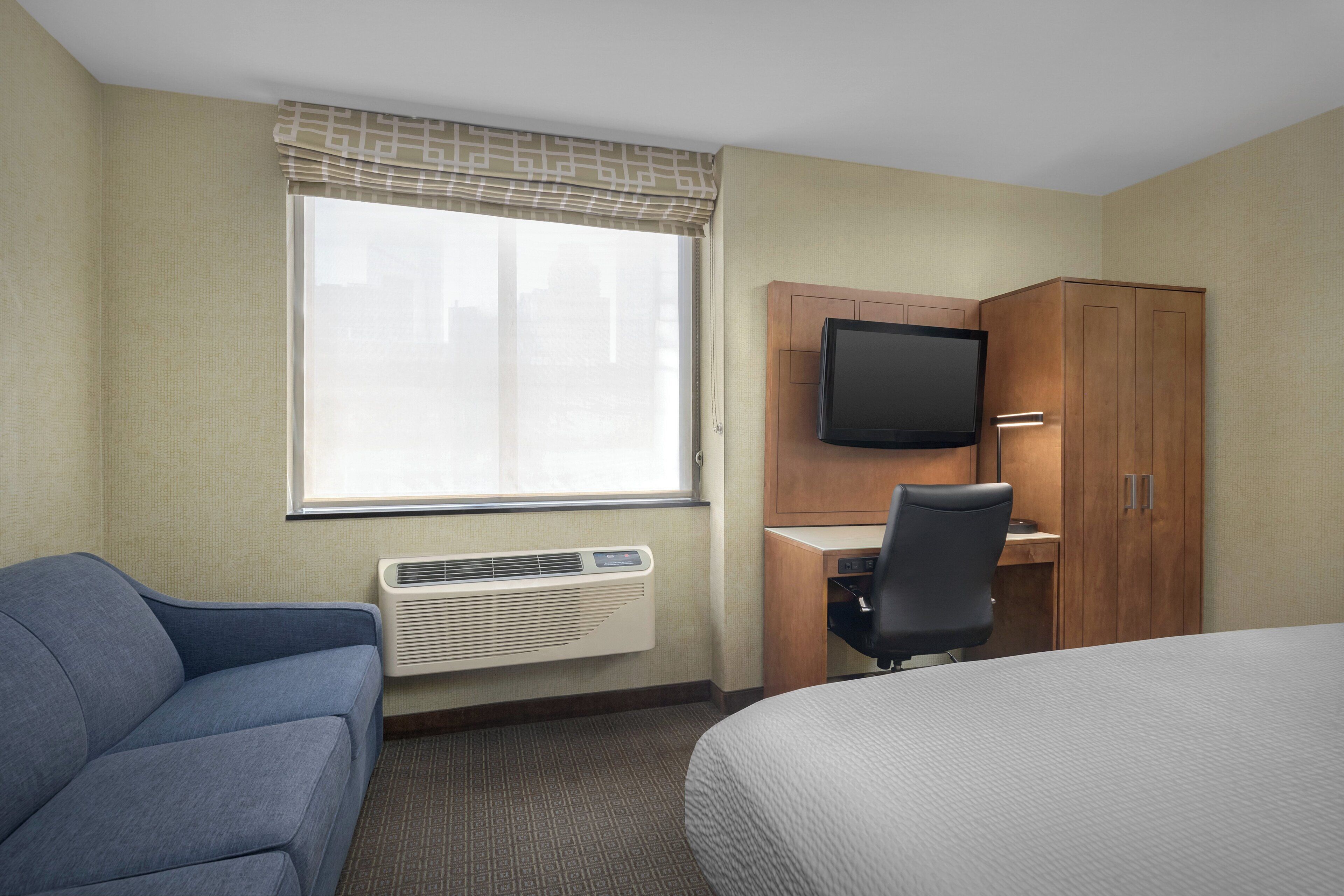 A hotel room with a bed, a desk with a television, a chair, and a sofa.