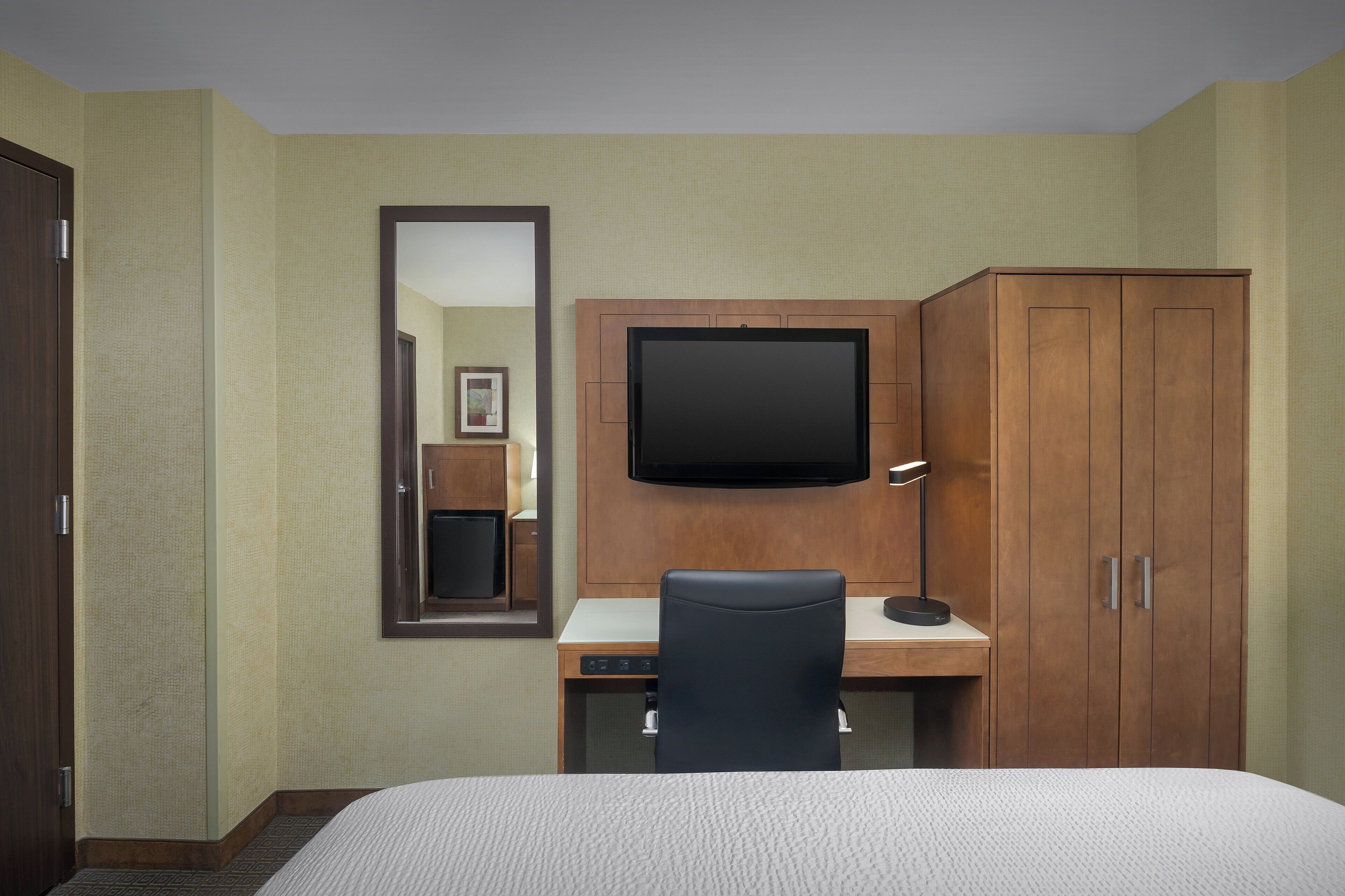 Quarto de hotel com cama, escrivaninha, cadeira, televisão e guarda-roupa.