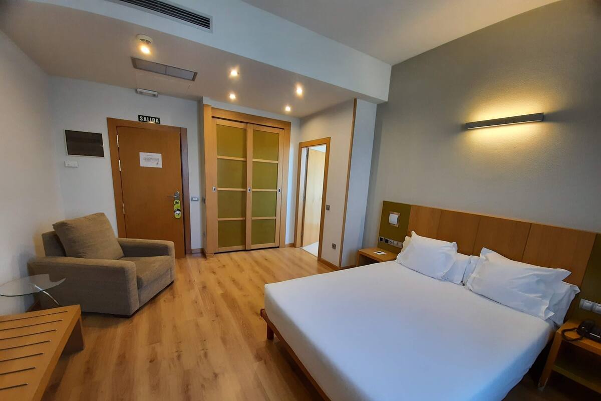 A hotel room with a large bed, a sofa, a small table, a nightstand, and a wardrobe with a sliding door.