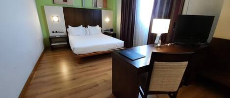 A hotel room with a bed, a desk with a chair, a TV, and artwork on the walls.