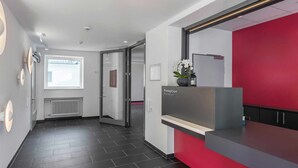 Een receptiezone met een rode accentmuur, een receptiebalie, een raam met zonwering, een radiator en een betegelde vloer.