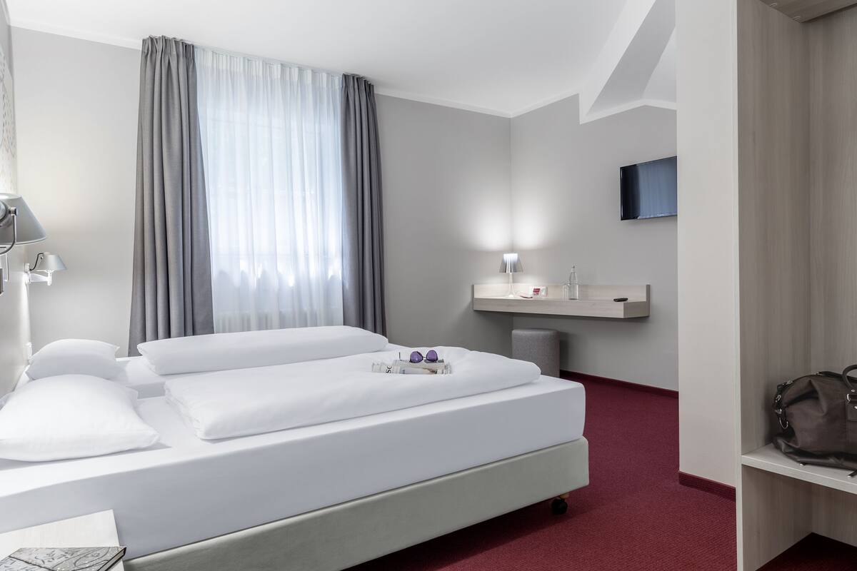 Een hotelkamer met een bed, nachtkastje met lamp, spiegel, televisie aan de wand en een vloer met rood tapijt.