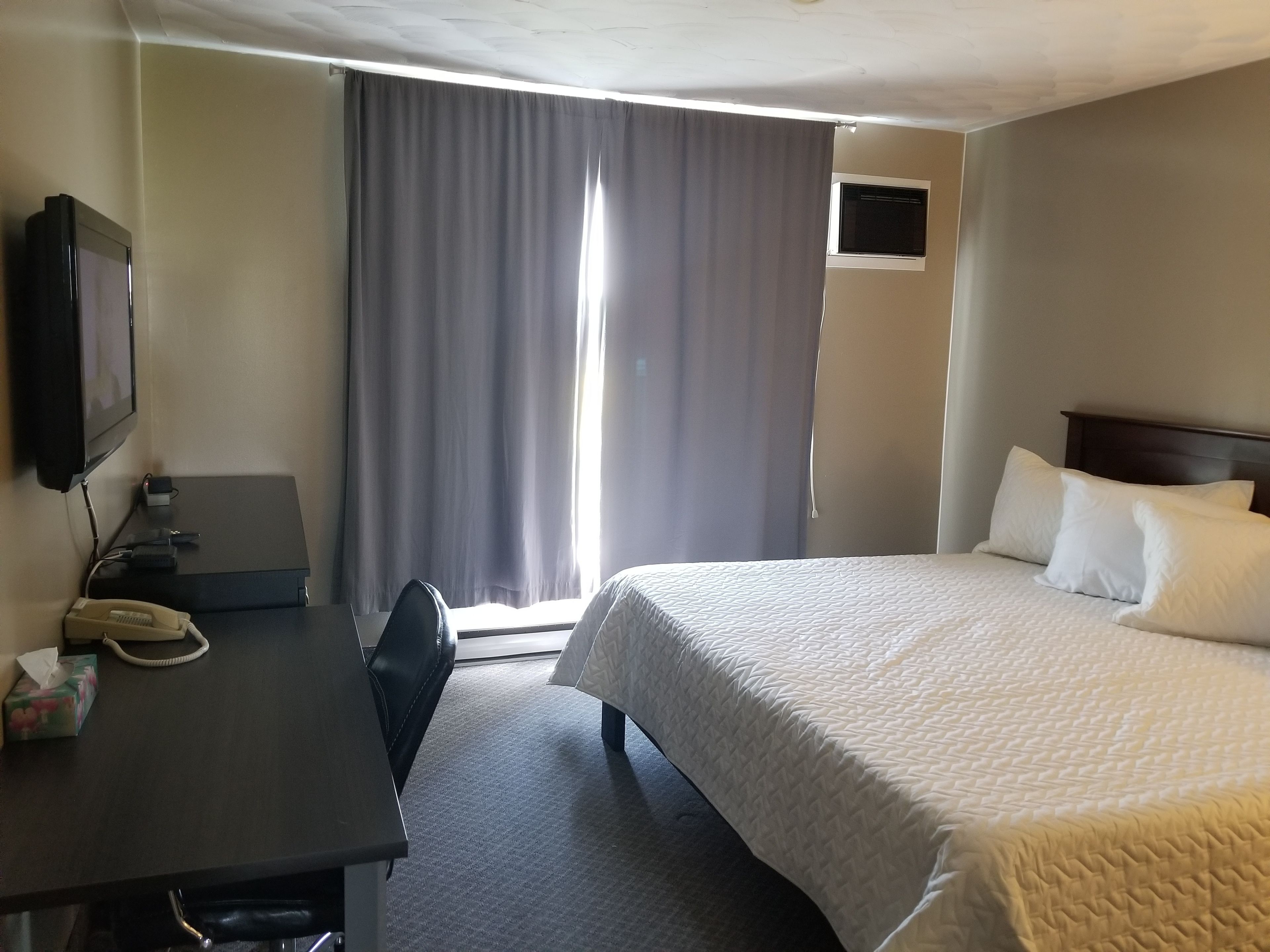 A hotel room with a bed, a desk with a telephone, a chair, a television mounted on the wall, and a window with curtains.