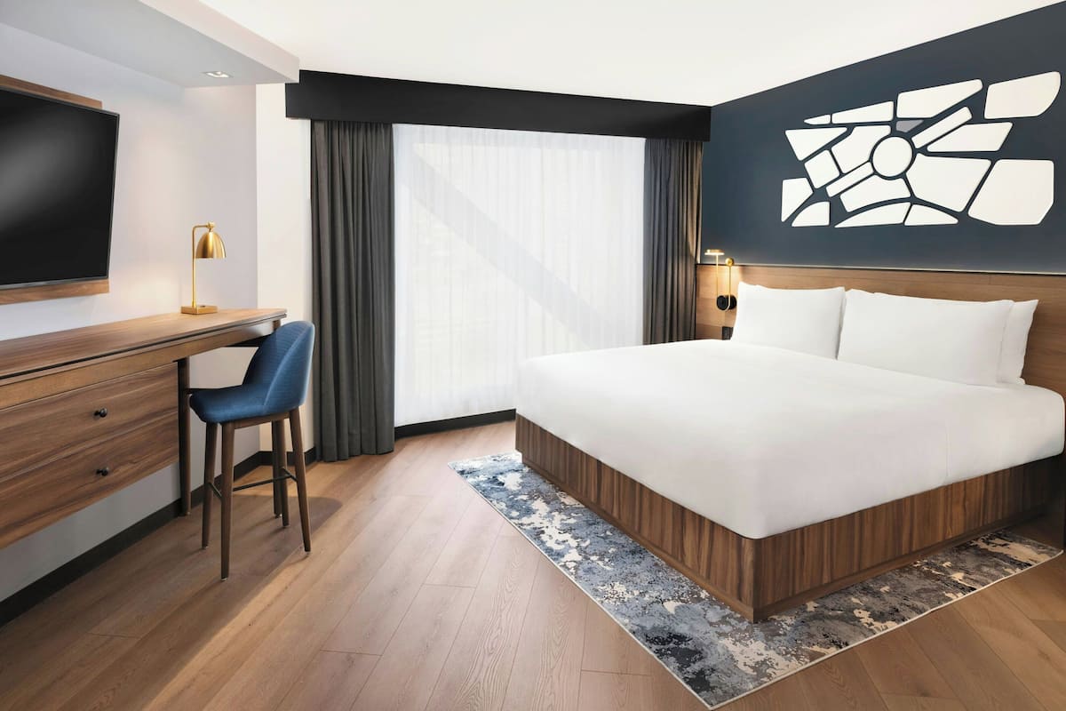 A modern hotel room with a large bed, a desk with a chair, a flat-screen TV, and a large window with curtains.