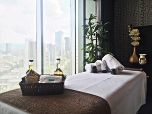 Habitación de hotel con un ventanal amplio que ofrece vista a la ciudad, una cama con sábana blanca, un cesto con botellas, una planta en maceta y un jarrón con flores secas.