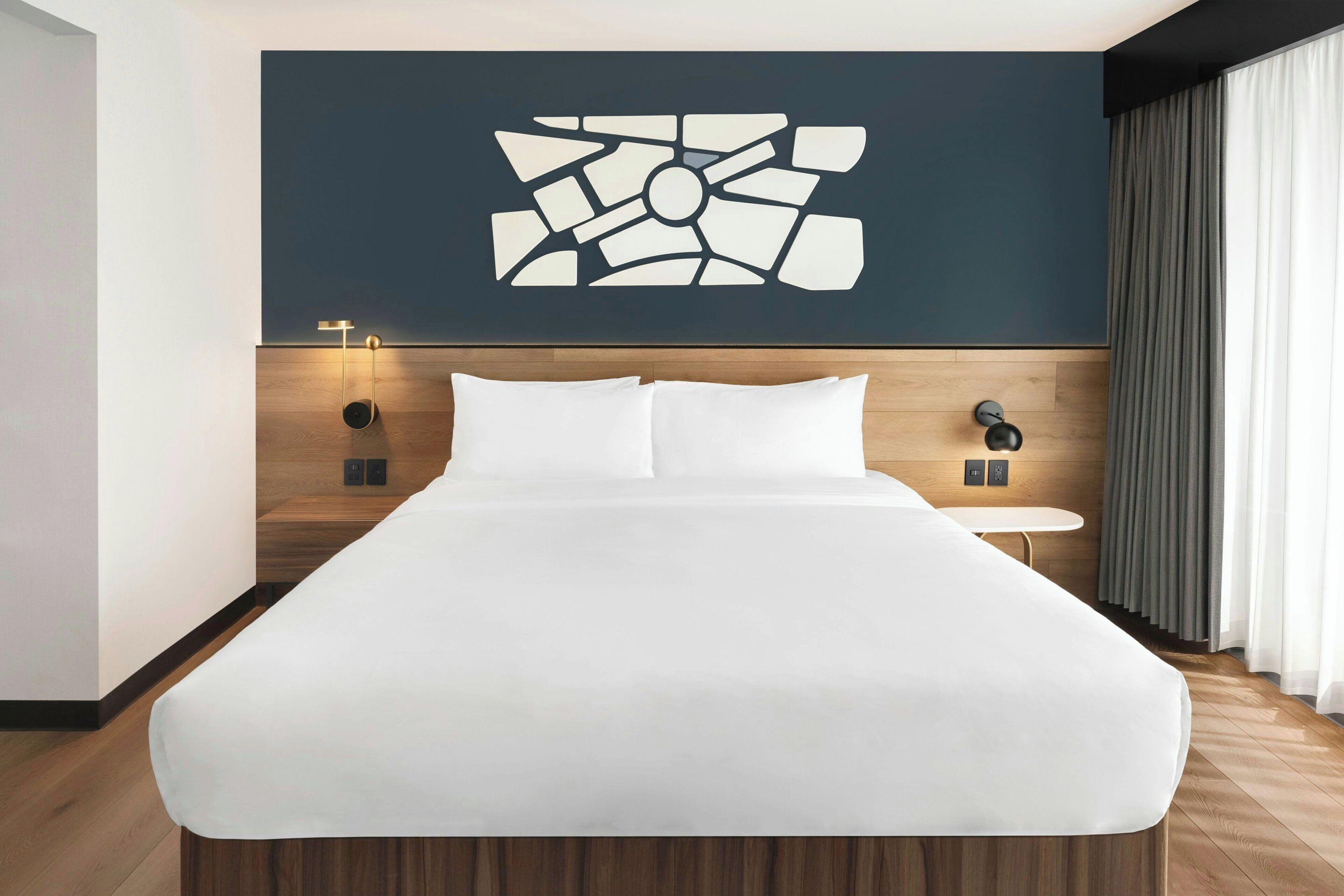 A modern hotel room with a large bed, a wall-mounted abstract art piece, a bedside table with a lamp, and a view of the outdoors through curtains.