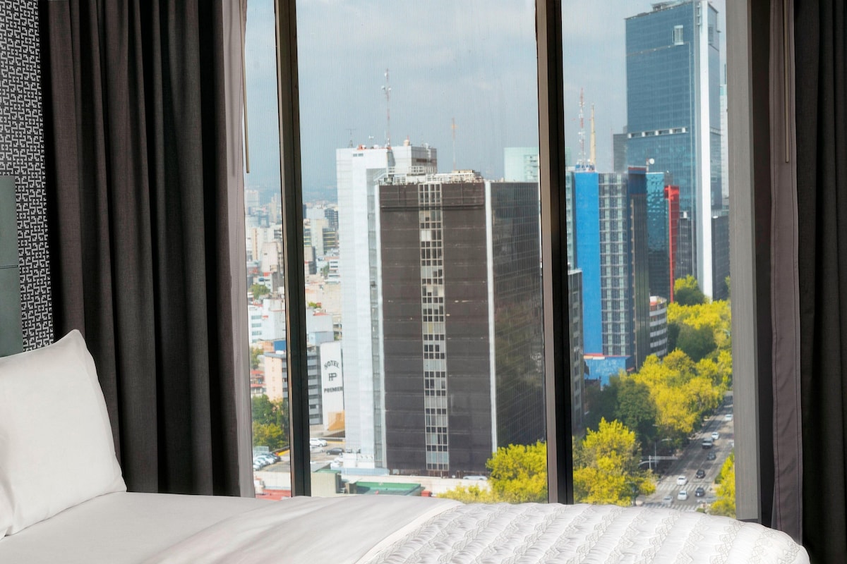 Een hotelkamer met een groot raam dat uitzicht biedt op een stadsgezicht met hoge gebouwen en een weg.