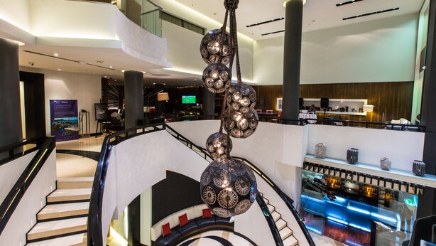 Een moderne hotelentree met een centrale trap, een bar en een scherm dat informatie toont.