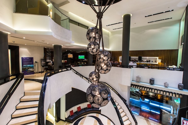 Un moderno vestíbulo de hotel con una escalera central, una zona de bar y una pantalla digital mostrando información.